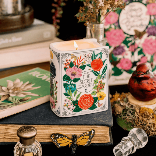 Lit Secret Garden book shaped candle styled on stacked books with floral décor in the background.