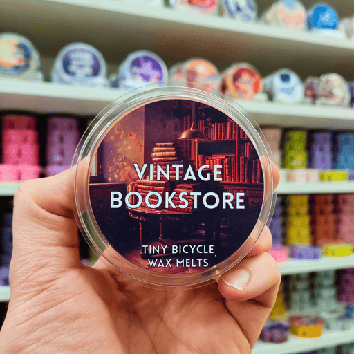 Vintage Bookstore wax melt pot held in hand with illustrated bookshop label, lifestyle image with wax melts on shelves in the background