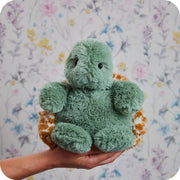 Warmies turtle plush held in hand with floral wallpaper background

