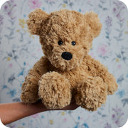 Warmies Brown Curly Bear plush held in hand against floral wallpaper

