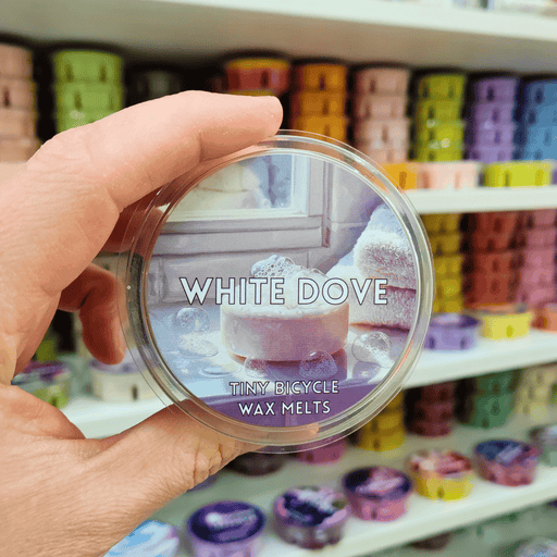 White Dove wax melt pot held in hand with soft pastel label, lifestyle image with colourful wax melts on shelves in the background