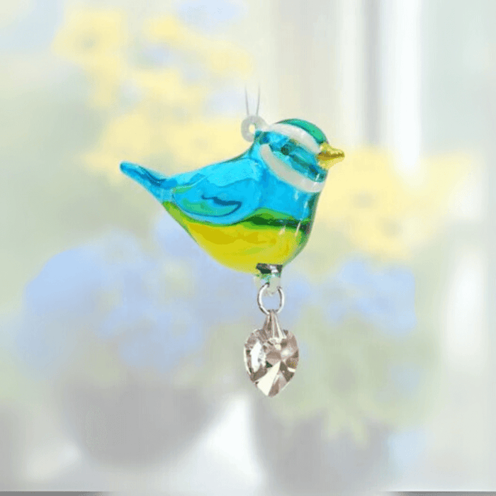 Handmade blue-and-yellow Blue Tit crystal suncatcher sparkling before soft flowers

