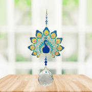 Wild Things Crystal Wonders majestic peacock crystal suncatcher hanging in a bright window, mirror-gold peacock motif in blue with 30mm crystal ball pendant.