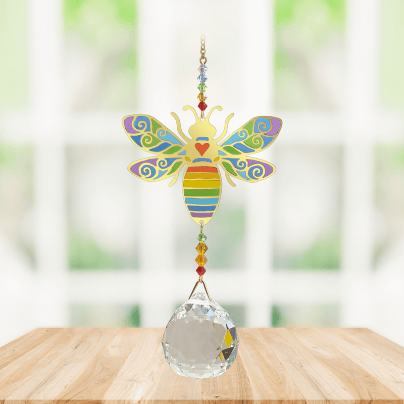 Wild Things Crystal Wonders rainbow bee crystal suncatcher hanging in a bright window, mirror-gold bee motif with rainbow detail and crystal ball pendant.