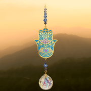 Hamsa Moonlight Crystal Suncatcher against sunrise hills – teal-lilac lotus hand, midnight-blue beads and 20 mm prism scattering colour.


