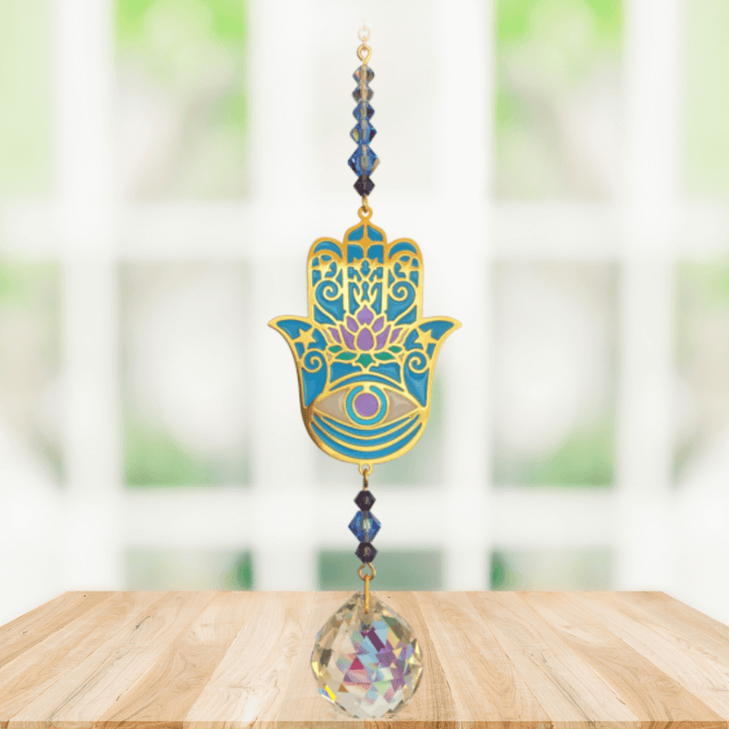 Hamsa Moonlight Crystal Suncatcher hanging in a bright window; ornate turquoise hand motif, sapphire beads and sparkling crystal ball ready to cast rainbows.
