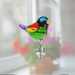 Handmade rainbow-coloured glass bird suncatcher sparkling before blurred blooms

