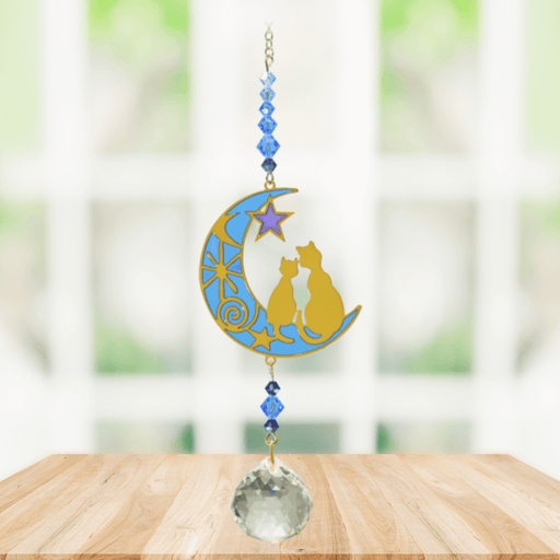 Two Cats Moonlight Crystal Suncatcher hanging in bright window; blue-gold crescent, twin cat silhouettes, midnight-blue beads and sparkling crystal ball.
