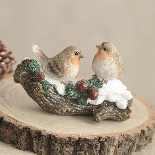 Lifestyle image of a robin duo ornament on a wooden slice, snowy branch and berries.