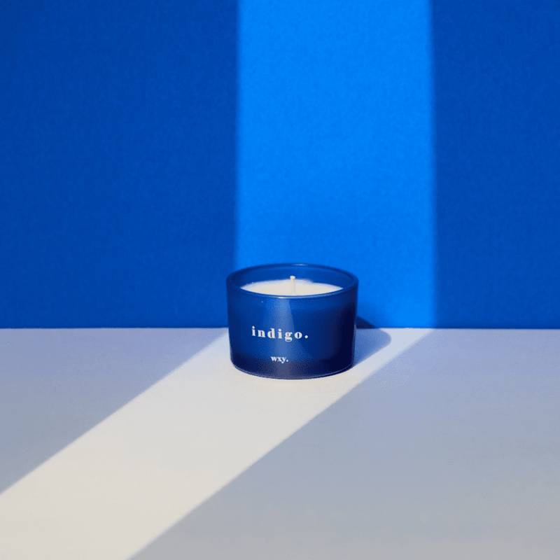 WXY indigo candle in blue glass styled in soft light against a vivid blue backdrop with long shadows.