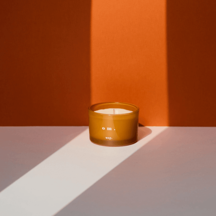 WXY om candle in amber glass styled in warm light against an orange backdrop with long shadows.