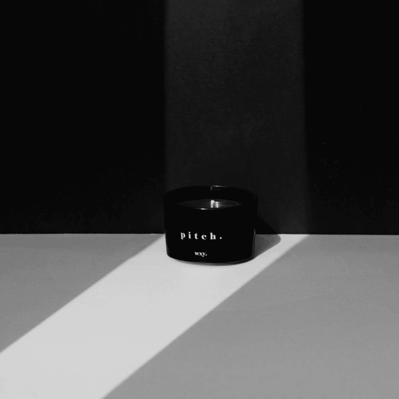 WXY pitch candle in black glass styled in soft light against a dark backdrop with long shadows.
