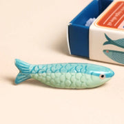 Mini blue-green ceramic sardine token outside the matchbox with sardine tin style box in background