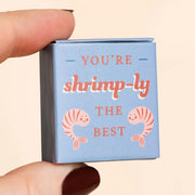 Hand holding the blue matchbox that reads You’re shrimp-ly the best with cute shrimp illustrations