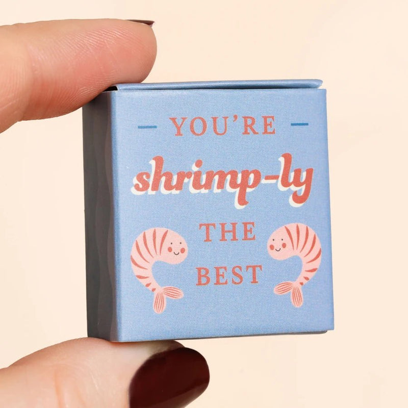 Hand holding the blue matchbox that reads You’re shrimp-ly the best with cute shrimp illustrations